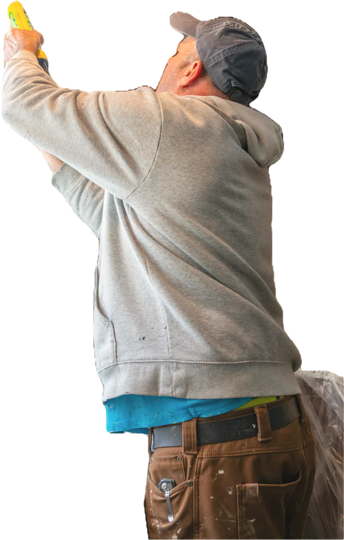A tradesman in a gray hoodie and cap, both arms up, sealing the edge of a range hood with a caulk gun.