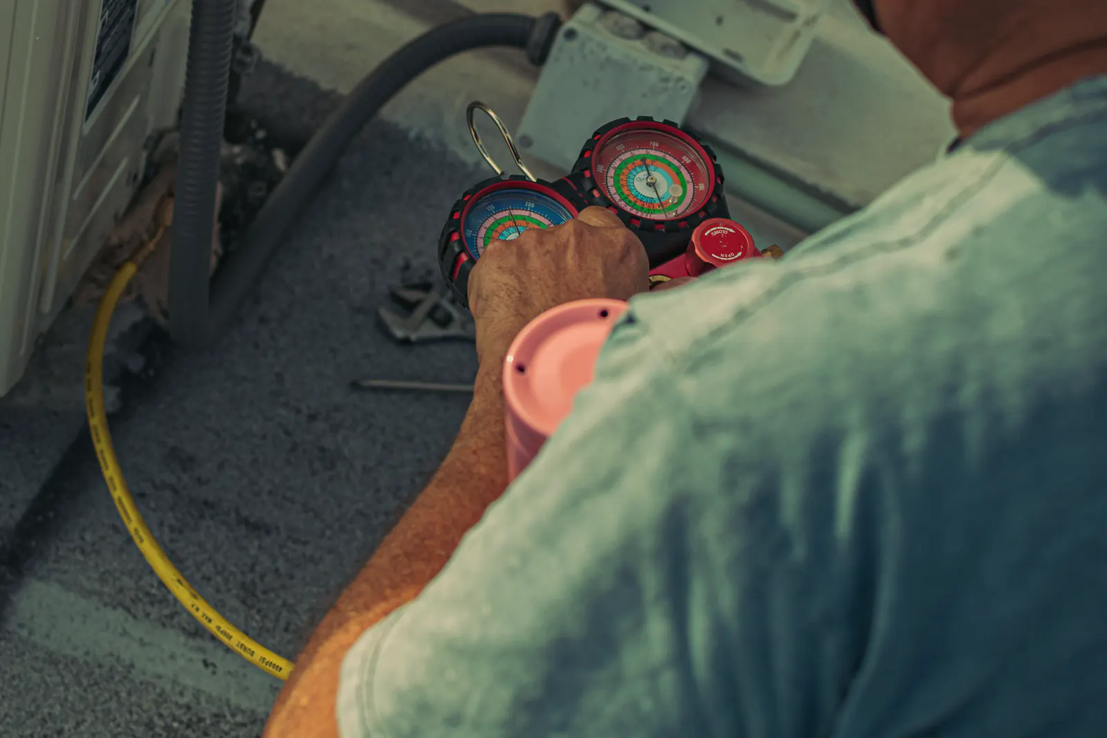 Technician servicing an outdoor air conditioning unit with refrigerant gauges and hoses.
