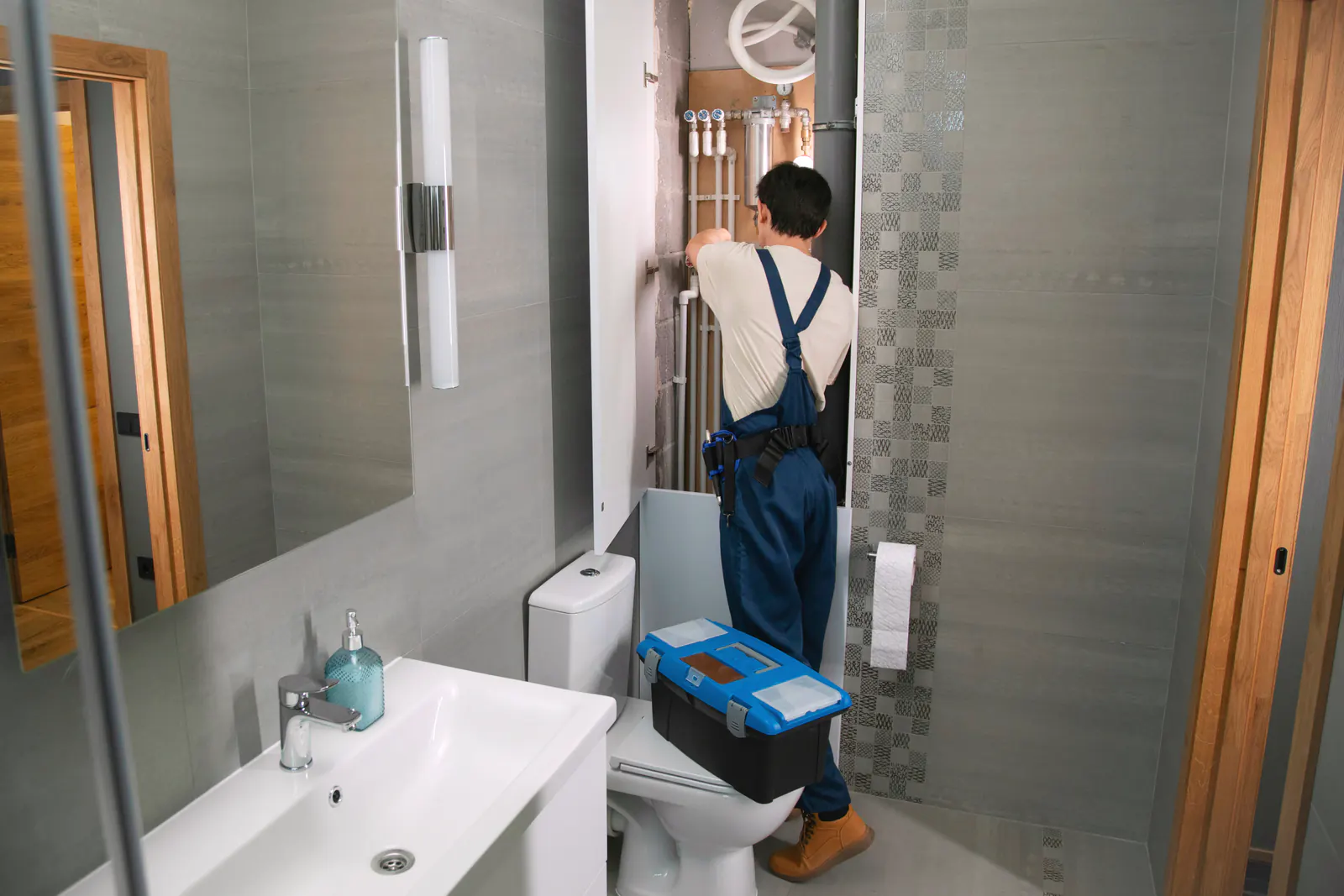 Plumber in blue overalls working on the plumbing in a bathroom.